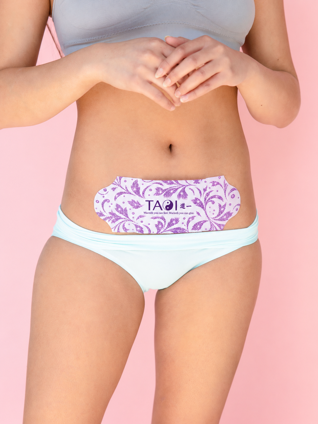 A model wearing light blue bikini bottoms and a light gray sports bra, with a purple and white damask-patterned TAOI abdominal patch applied to her lower abdomen, set against a solid pink background.