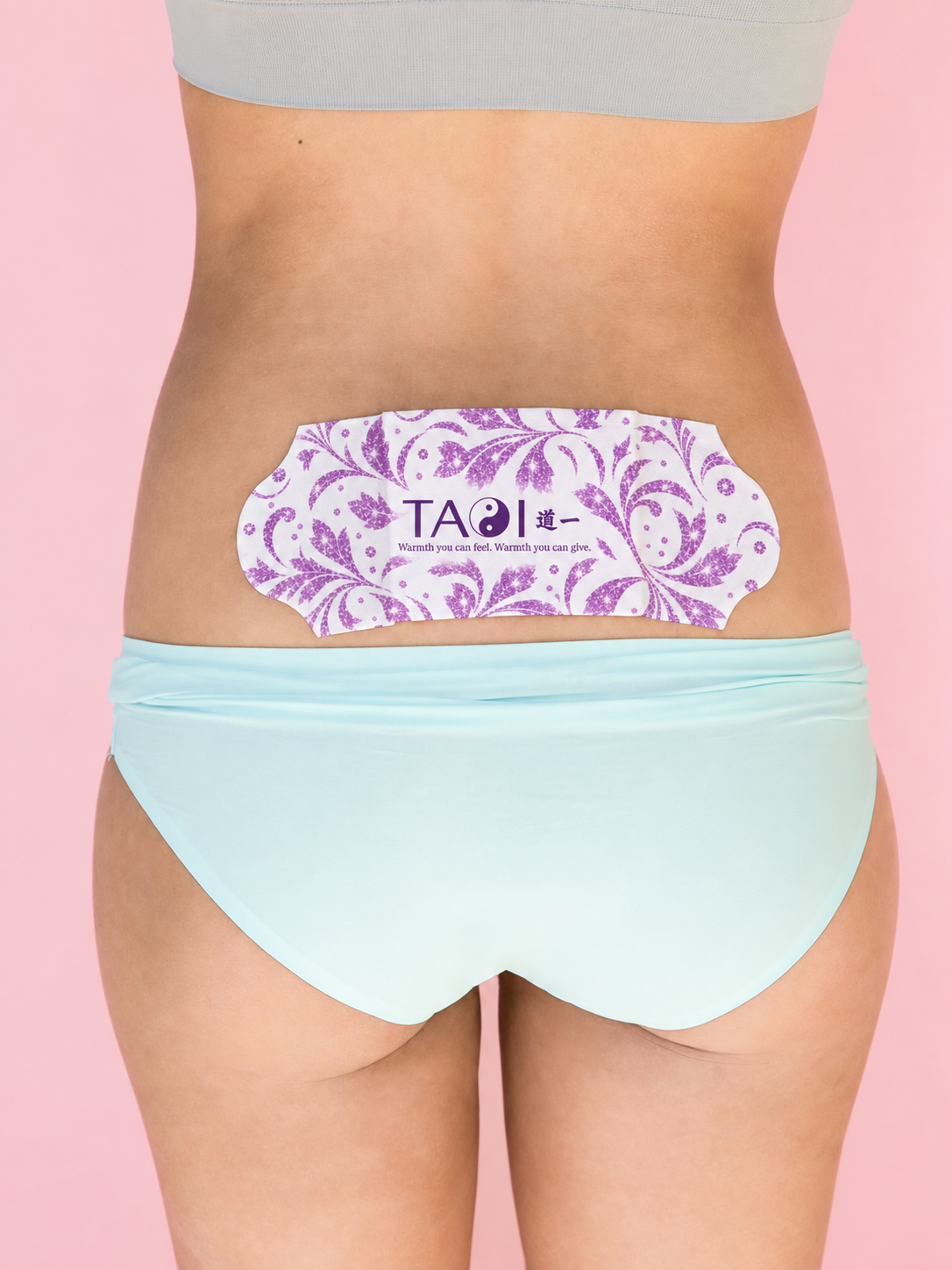 A close-up of a person's lower back wearing a light blue bikini bottom and a white adhesive patch with purple paisley patterns and the brand name 'TAO' in the center, set against a solid pink background.